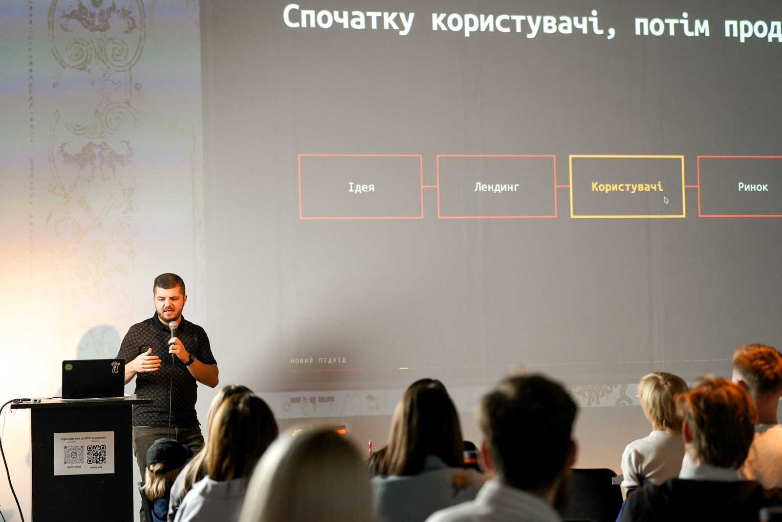 Kraków Tech Mixer #2: Ship It, 3 вересня 2026 року, HEVRE. Фото: ВУС. Спікер з мікрофоном біля трибуни розповідає на тлі слайда, перед ним глядачі на Kraków Tech Mixer #2